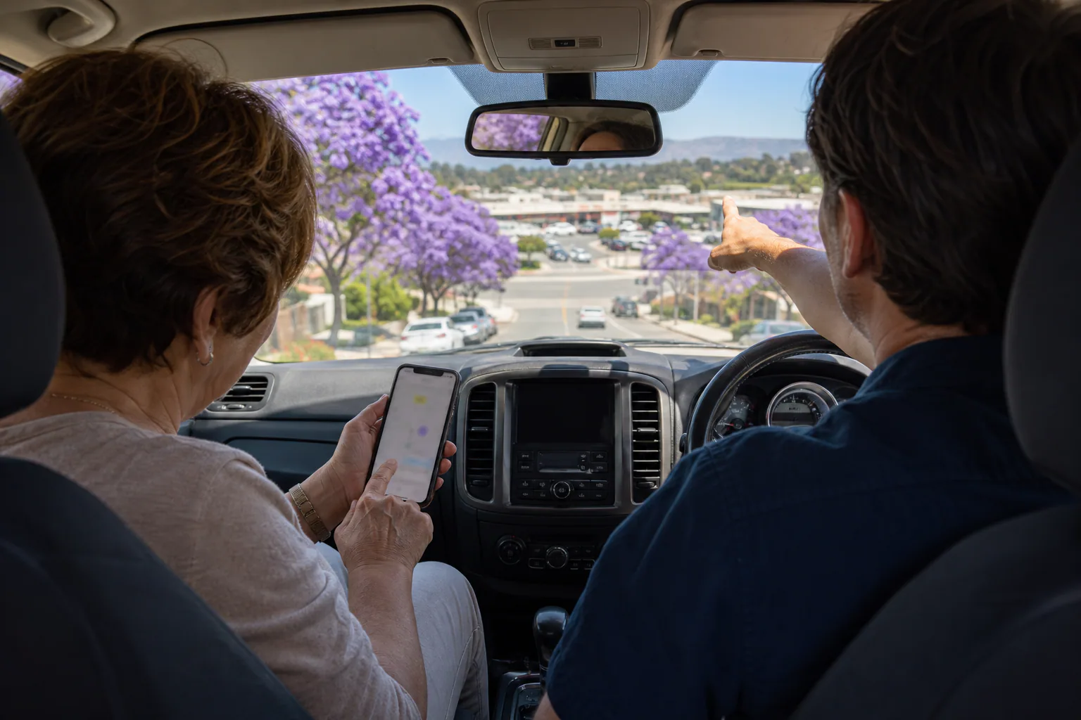 Woman in the passenger seat checks a map app while her son points toward a distant shopping strip from a parked car