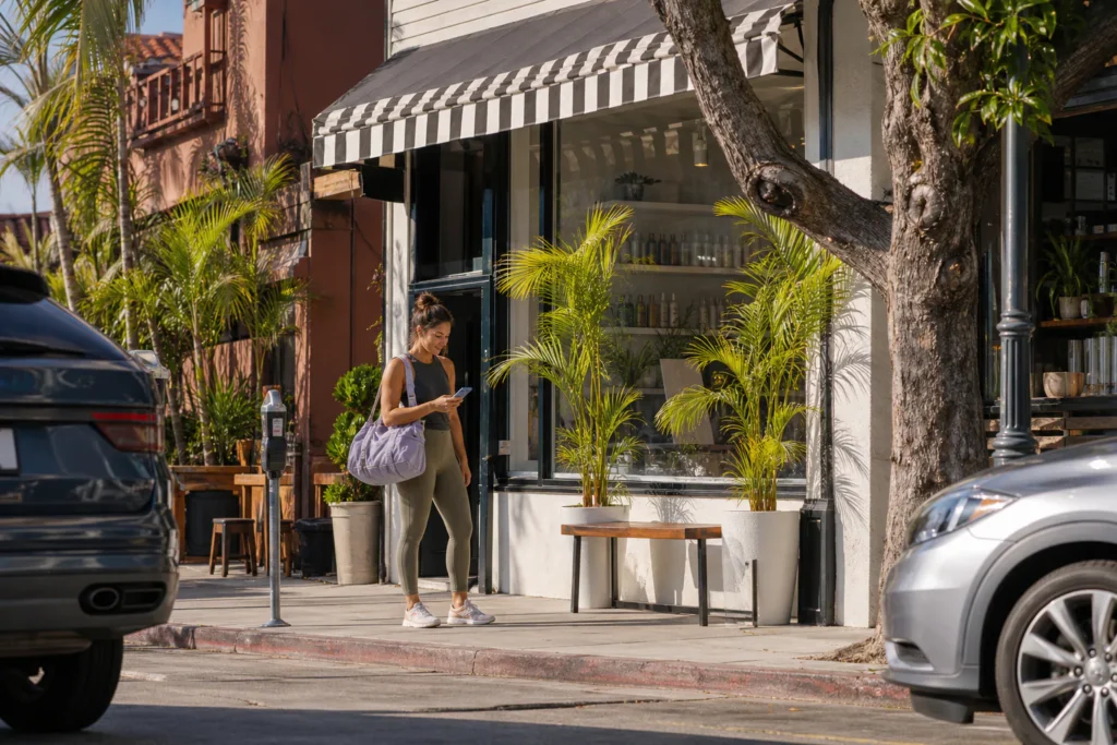 Woman with a gym bag checks her phone outside a small neighborhood hair salon, looking for the right storefront on a sunny street